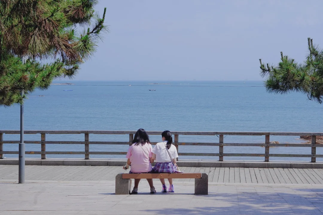 Two girls sit on a bench overlooking the ocean.