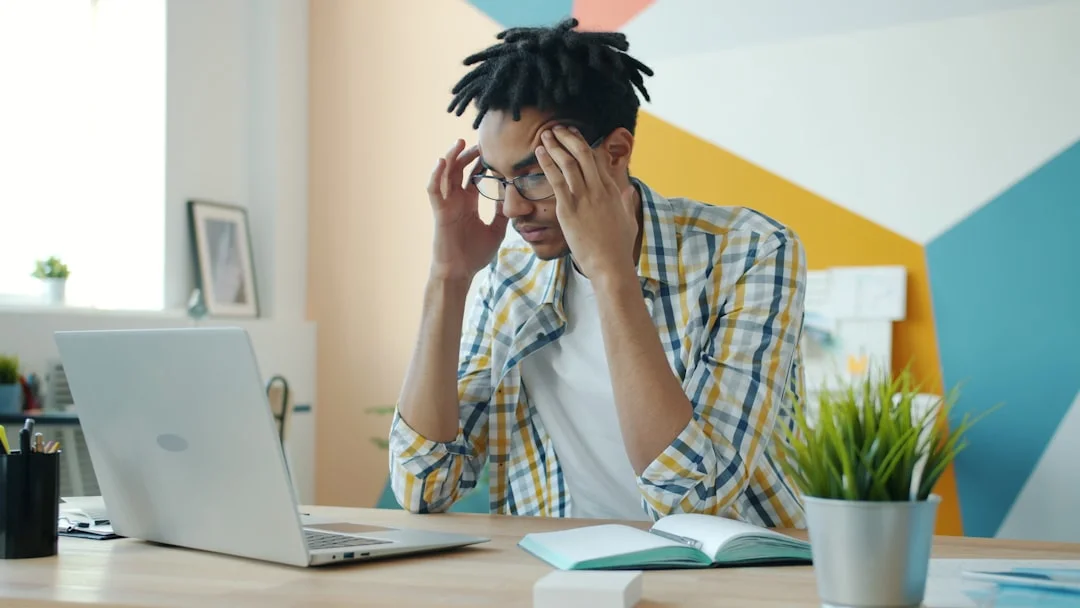 Man with dreadlocks holding head at desk with laptop