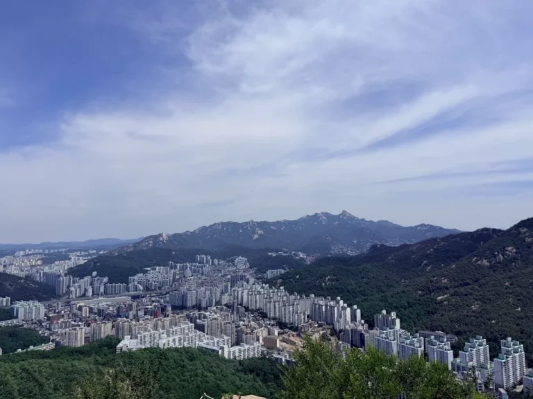 Urban landscape with buildings and mountains under a cloudy sky.